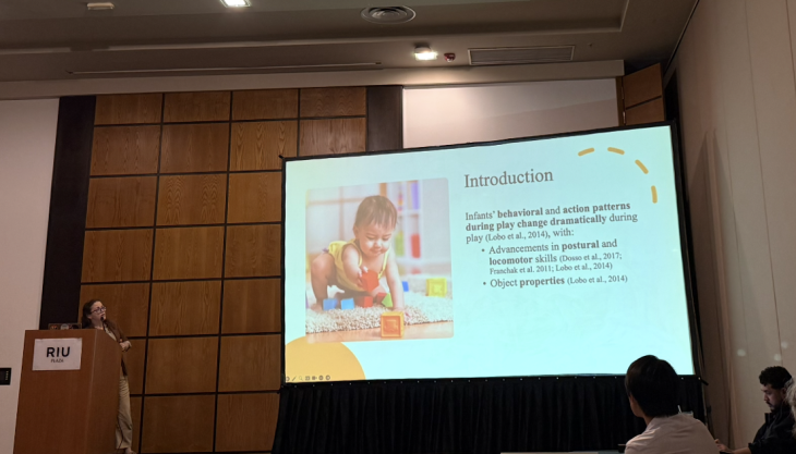 A presenter stands at a podium delivering a research presentation while a large screen displays an introductory slide featuring a child playing and text about infant behavioral patterns. Audience members watch from the conference room.